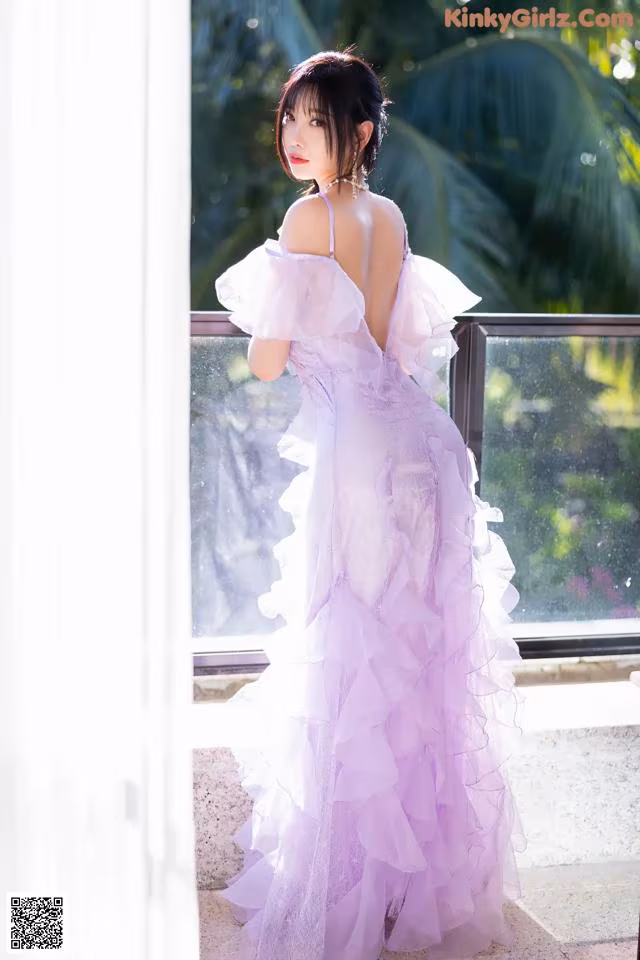 A woman in a purple dress standing by a window.