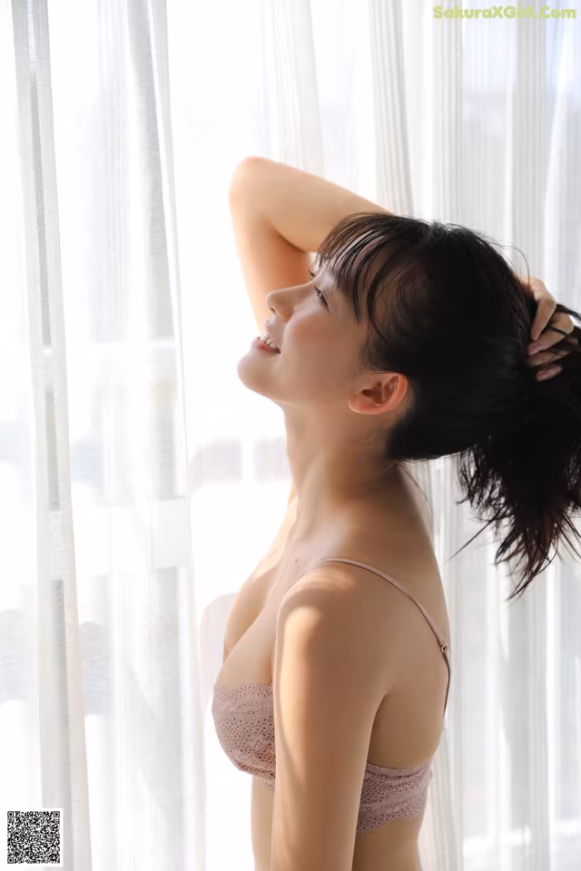 A woman in a pink bra standing by a window.